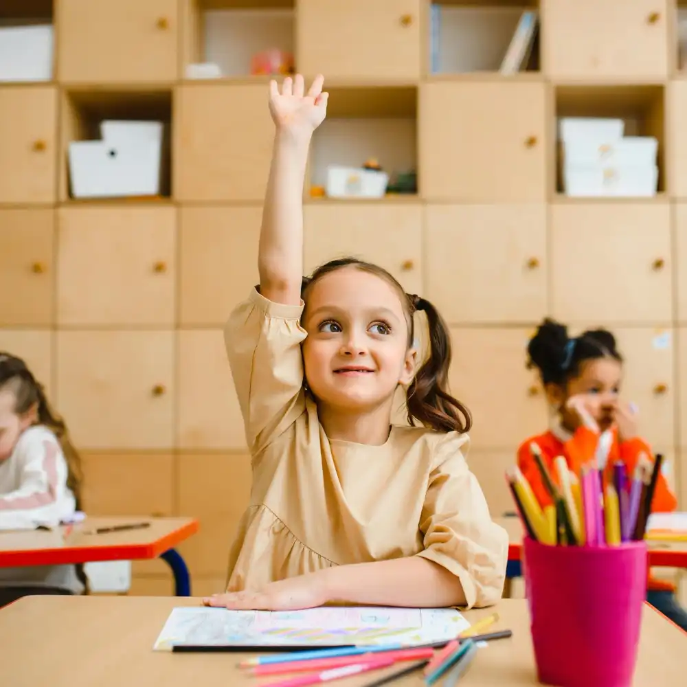 Smiling girl raising her hand confidently in a classroom during an art or learning activity.