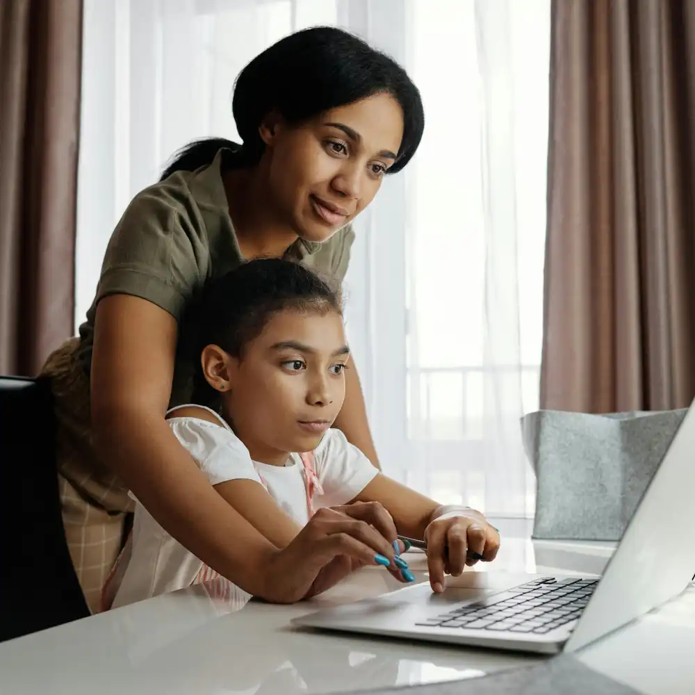 Teacher helping a young child with online learning on a laptop at home, both focused on the screen.