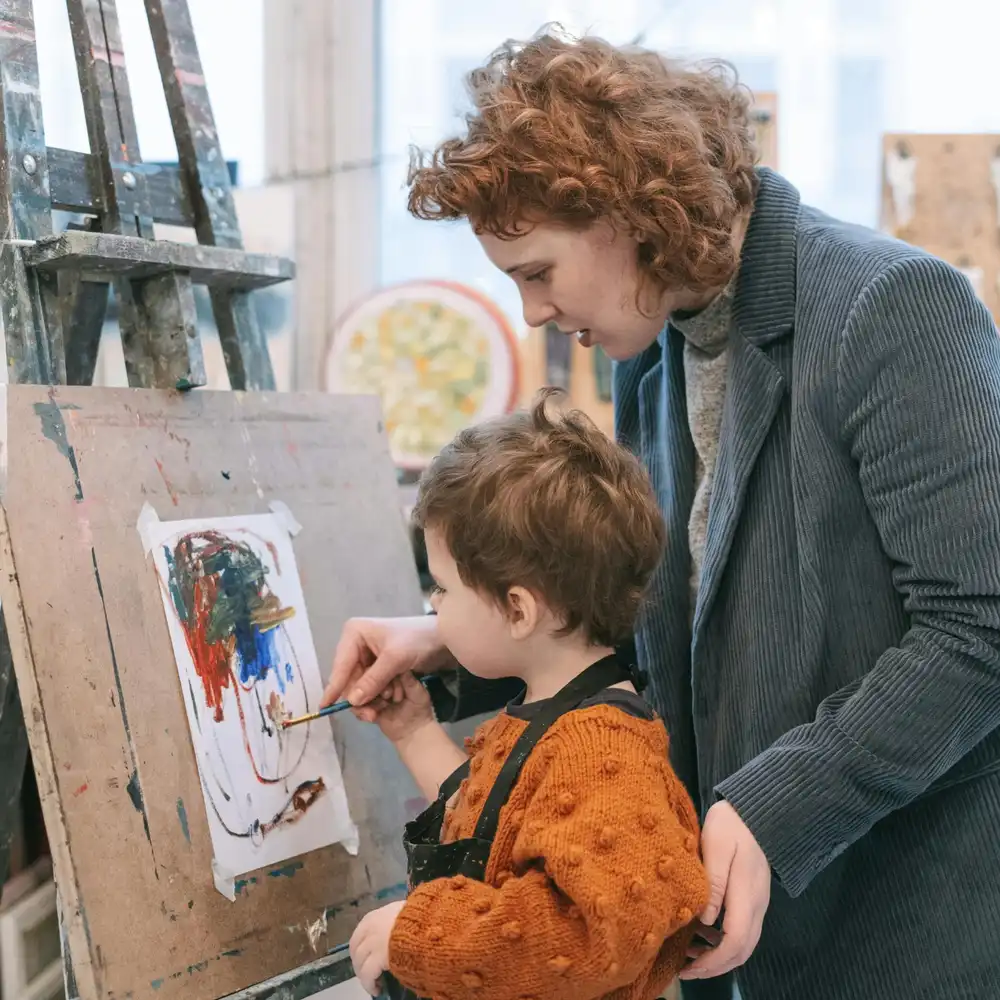 Teacher guiding a young child painting on an easel during an art lesson in a classroom studio.
