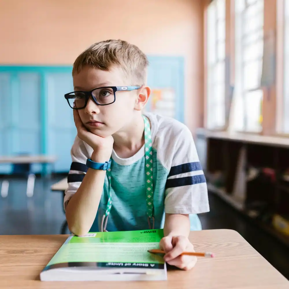 Thoughtful student sitting at a desk in a classroom, looking away while holding a pencil and notebook.